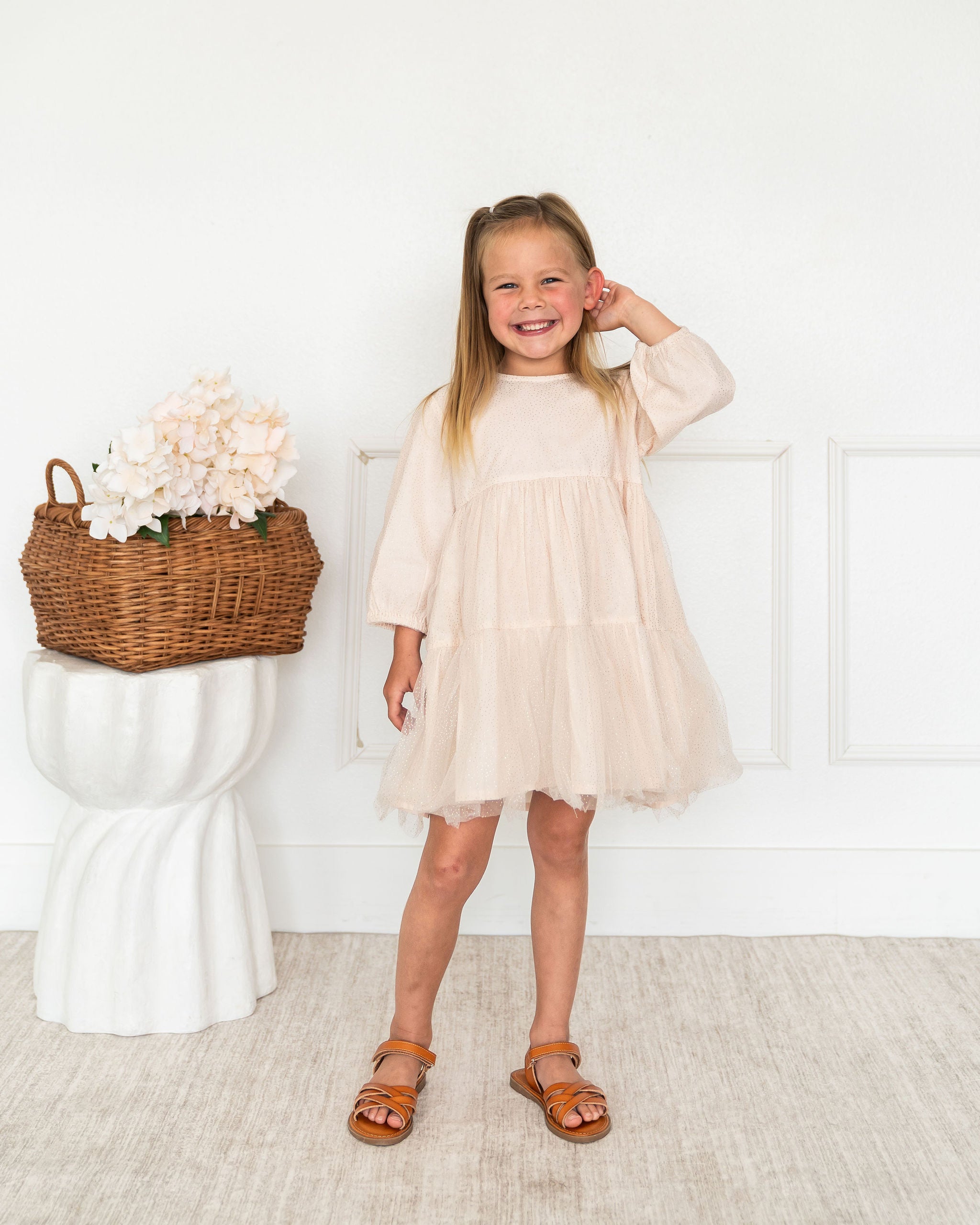 A young girl with blonde hair stands smiling in a room, wearing a light beige tiered tulle dress with three-quarter sleeves and brown sandals. To her left is a white pedestal holding a wicker basket filled with pale pink flowers.
