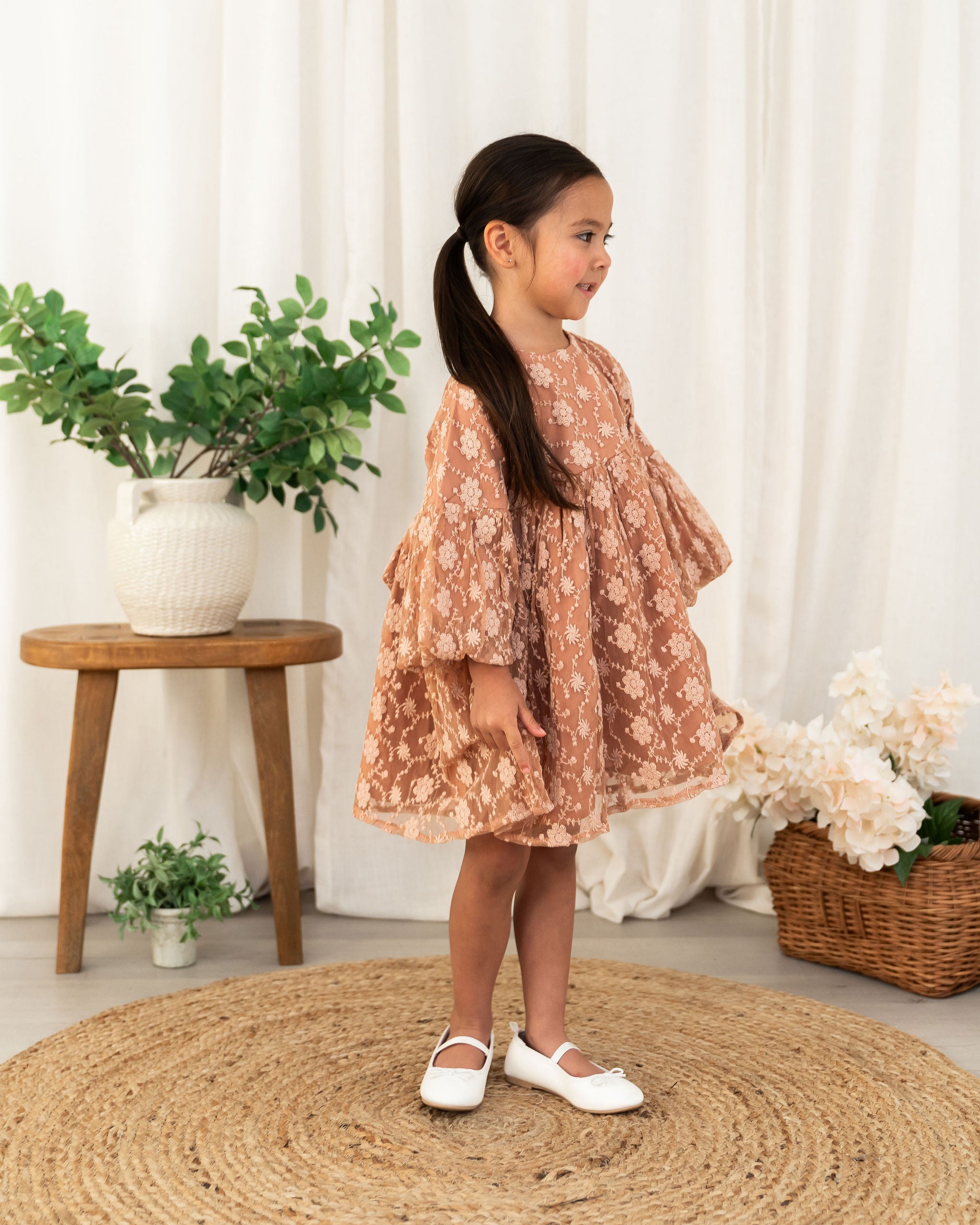 A young girl with a ponytail stands in profile wearing a dusty rose tiered organic lace dress with a beige floral pattern, styled with white Mary Jane shoes on a round jute rug in a bright, neutral room.