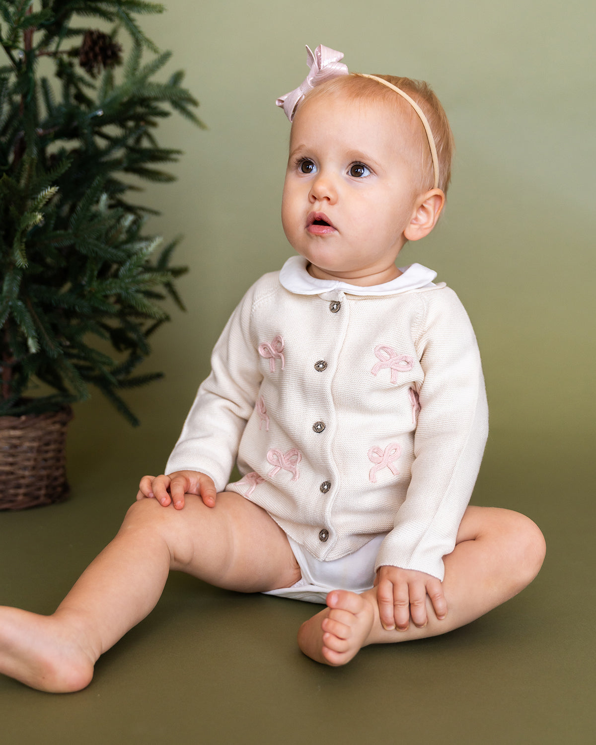 Viverano Organics baby cardigan in cream with pink bow embroidery worn by a seated baby girl