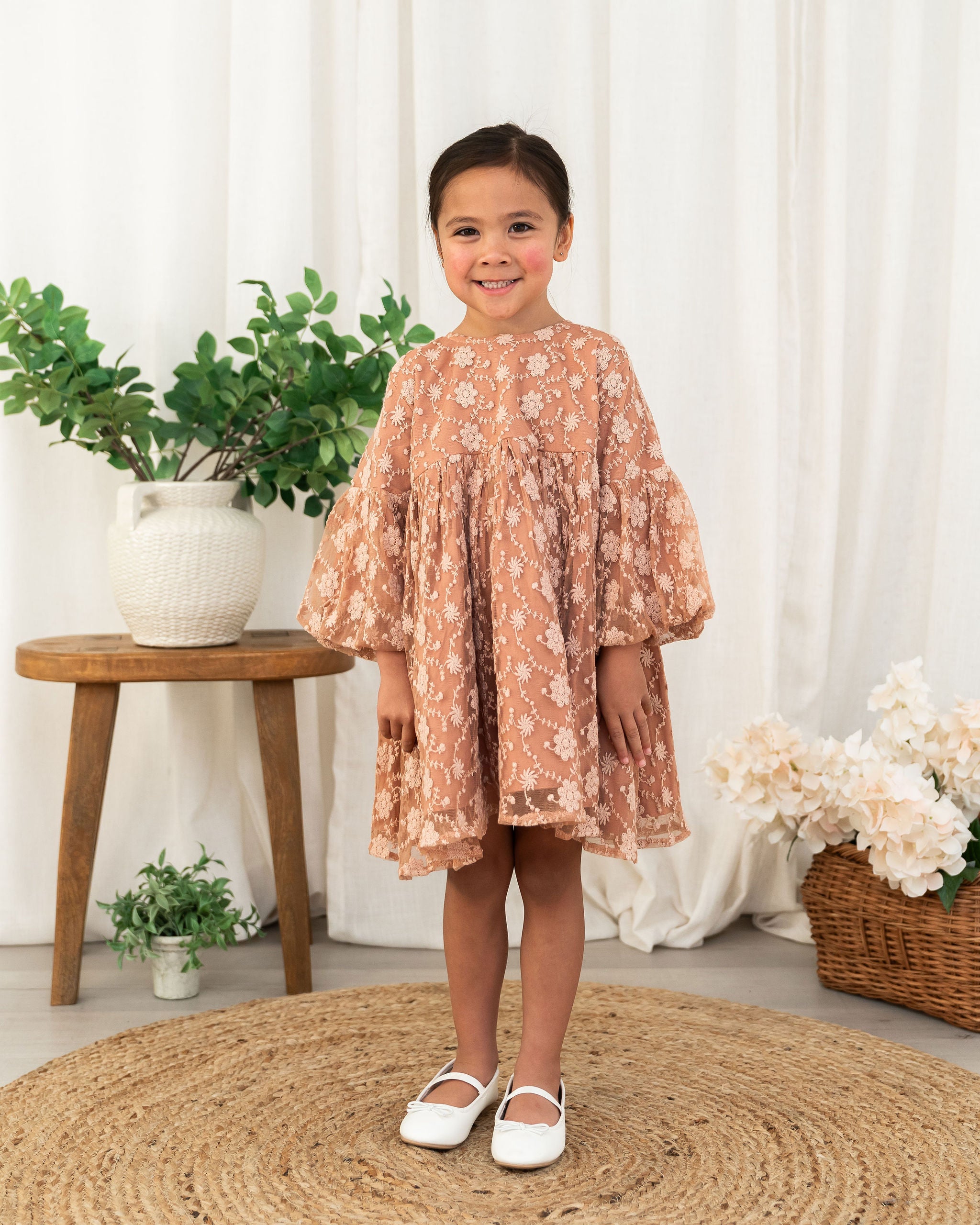 A toddler girl stands smiling in a dusty rose organic lace dress with white floral embroidery, paired with white Mary Jane shoes, set against a soft white curtain backdrop with decorative plants and a woven rug.